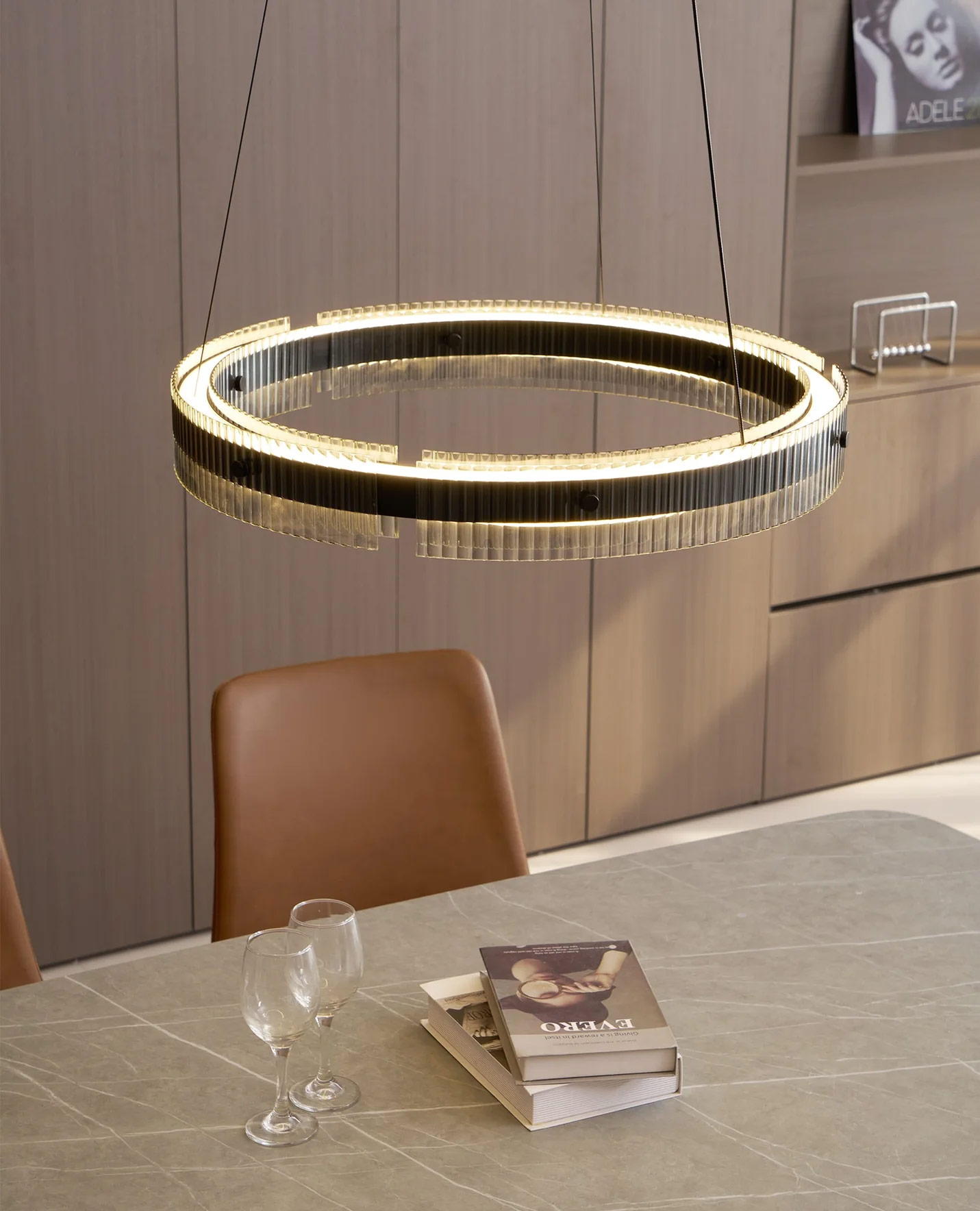 Contemporary double-ring pendant chandelier in black aluminium and fluted glass emitting soft 3000K LED light over a dining table with leather chairs and books.